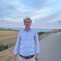 Young man in a light-blue shirt standing on a rural gravel road with fields and a fence under a blue sky behind him.